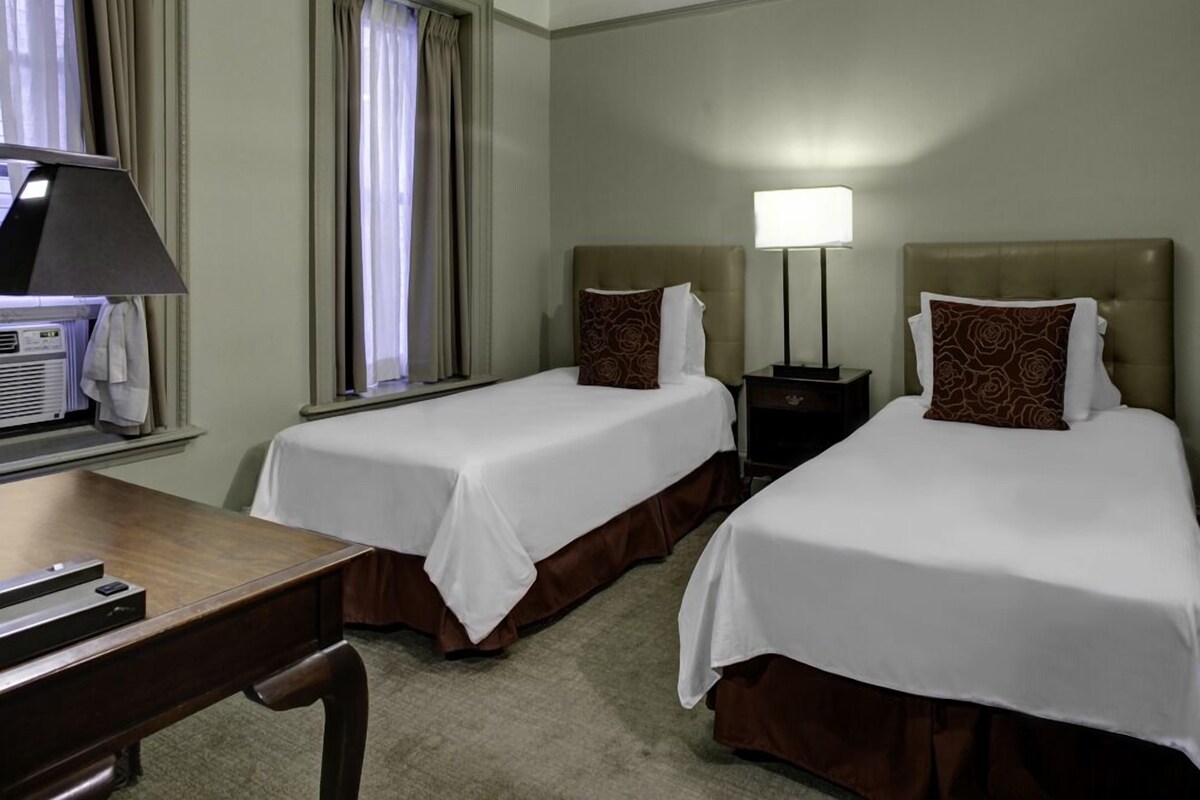 A hotel room with two beds, a desk, a lamp, and an air conditioning unit.