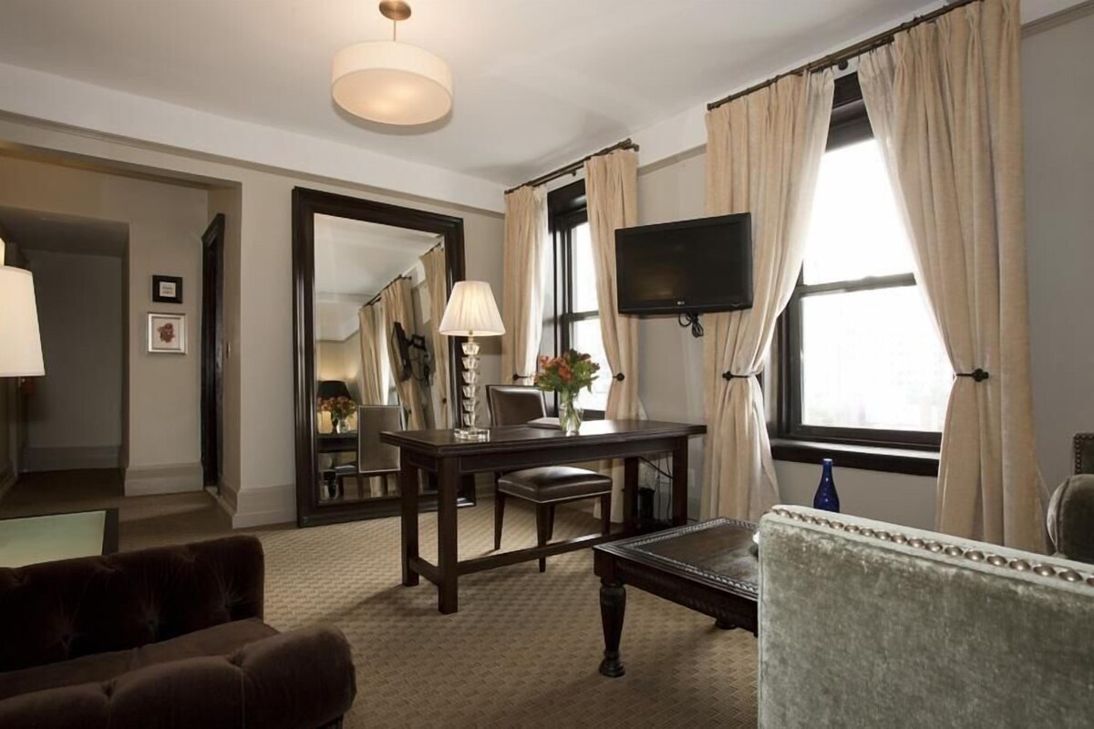 A hotel room with a large mirror, a TV mounted on the wall, a dining table with chairs, a sofa, and a window with curtains.