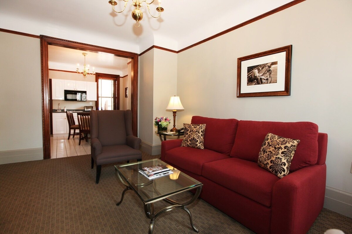 A living room with a red sofa, a glass coffee table, and a brown armchair. There is a dining area visible through an open doorway.