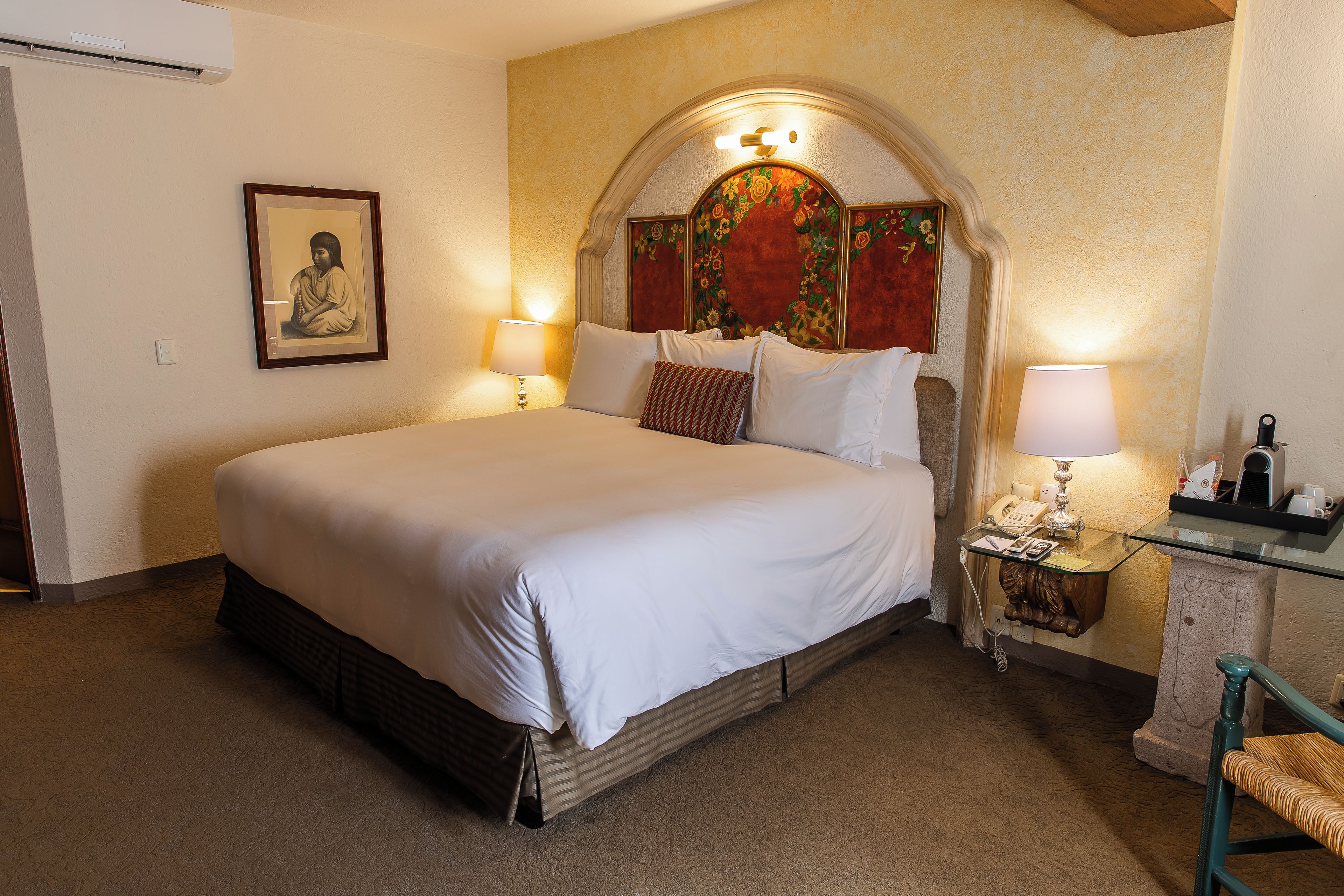 A neatly made bed with white linens, a wooden headboard with a floral design, two bedside lamps, and a small table with a telephone and a cup holder.
