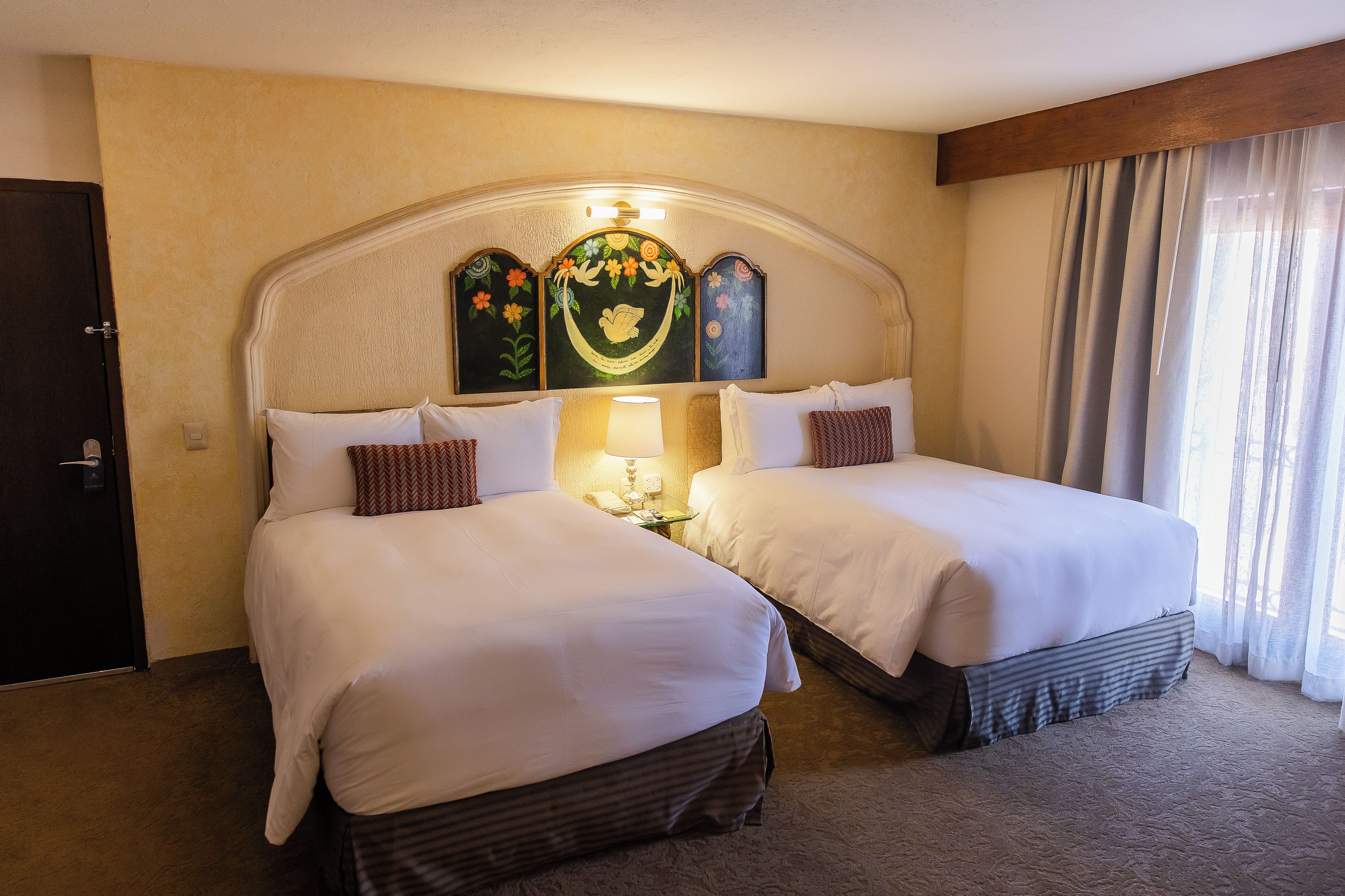 Habitación de hotel con dos camas, cabecera de madera, mesita de noche con lámpara y ventana con cortinas.