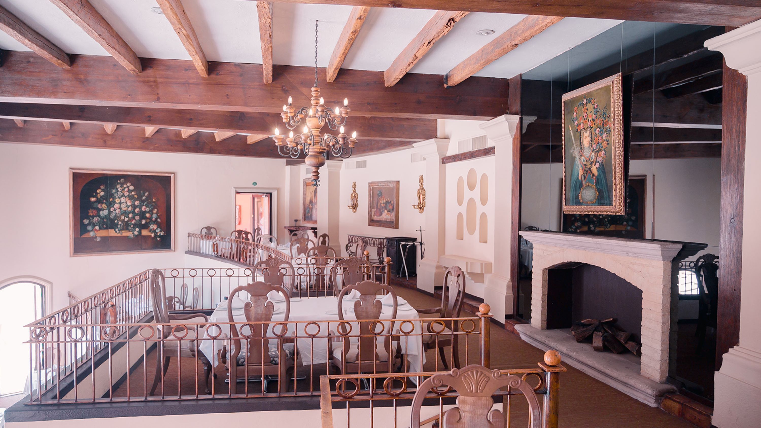 A dining area with a high ceiling, wooden beams, a chandelier, and a fireplace.