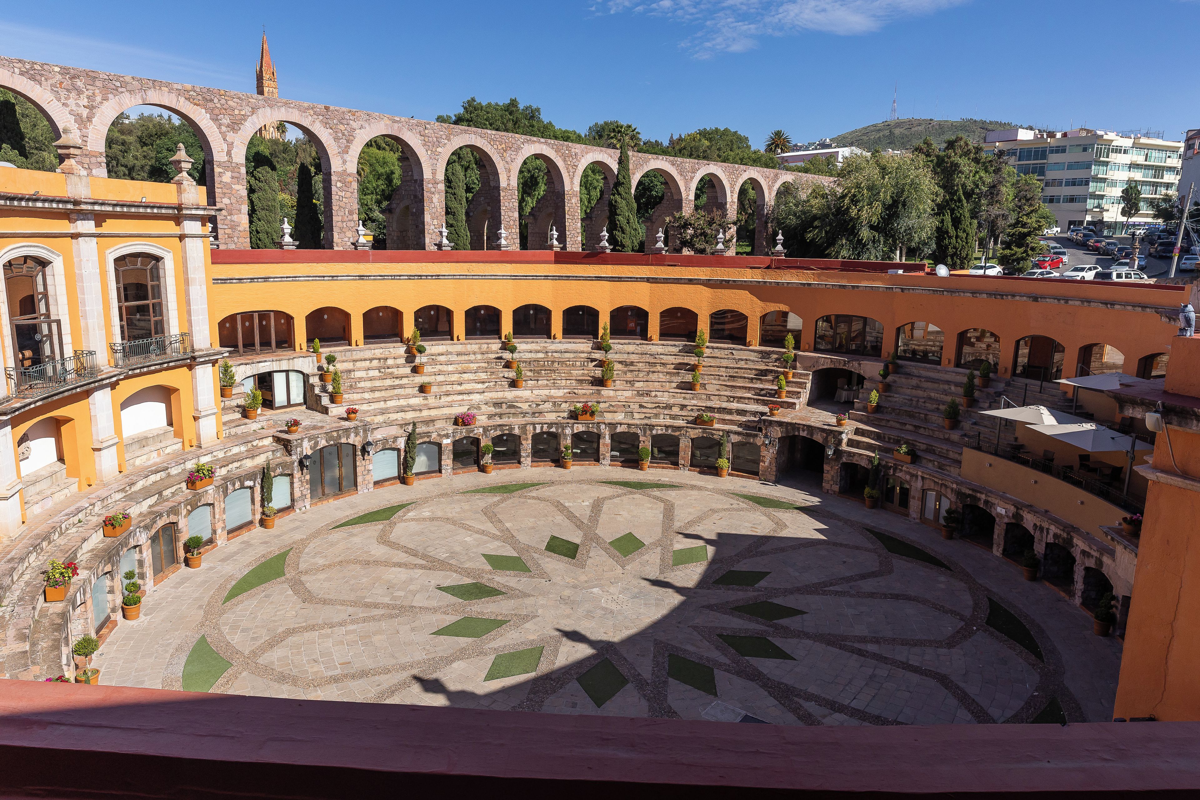 A historic courtyard with a central open space surrounded by arches, potted plants, and a tiled floor.