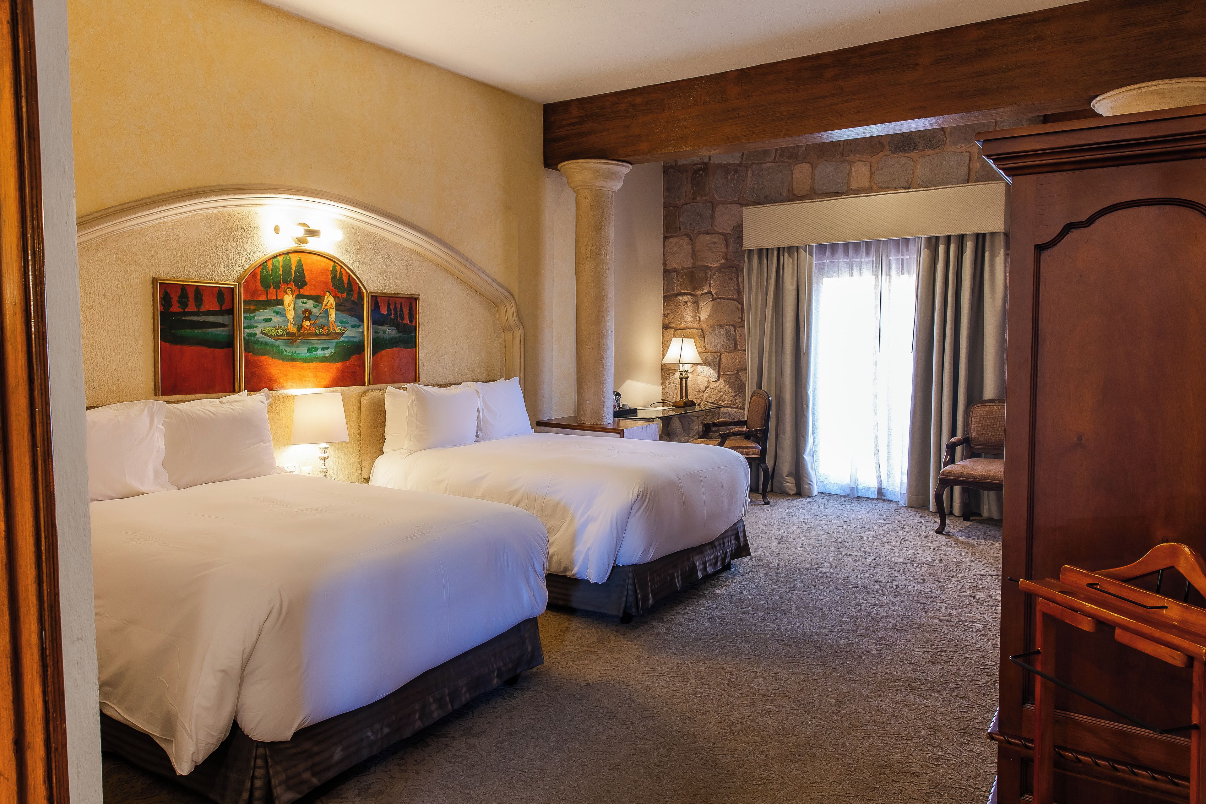 A hotel room with two beds, a stone wall, a large painting, a lamp, and a chair.