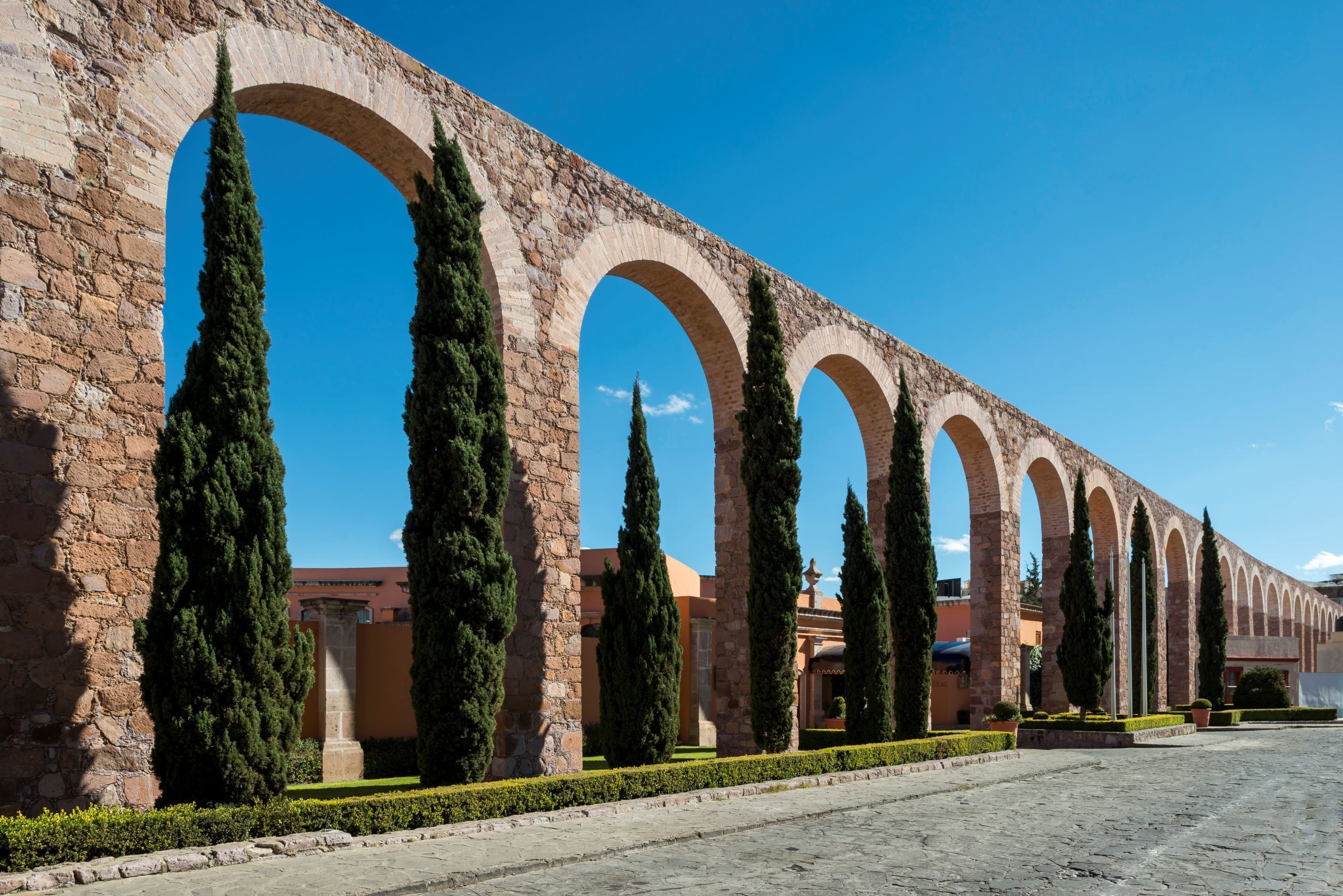 Una serie de arcos con un muro de piedra y altos cipreses en un patio.