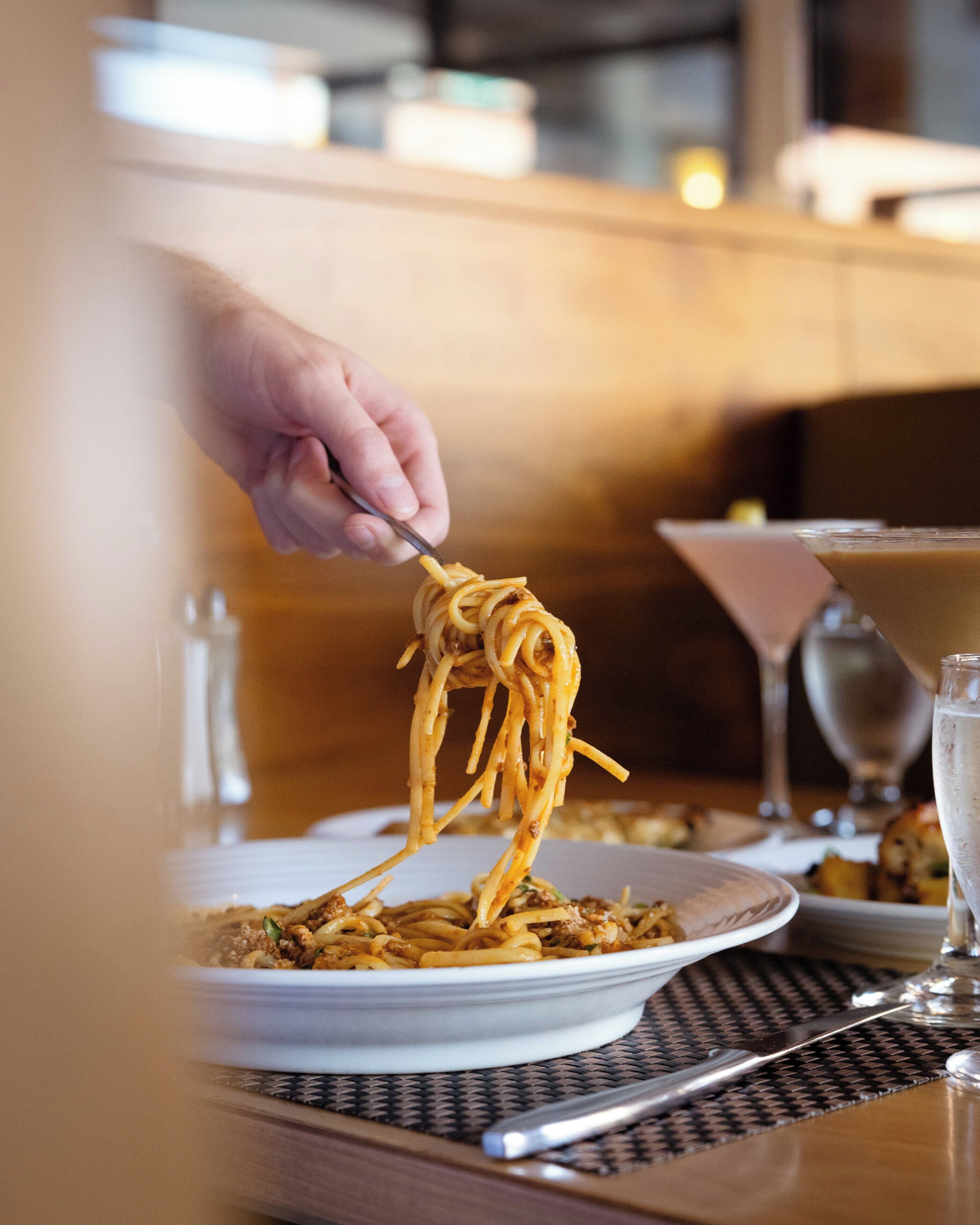 A dining table with a plate of spaghetti, a fork, a martini glass, and a wine glass.