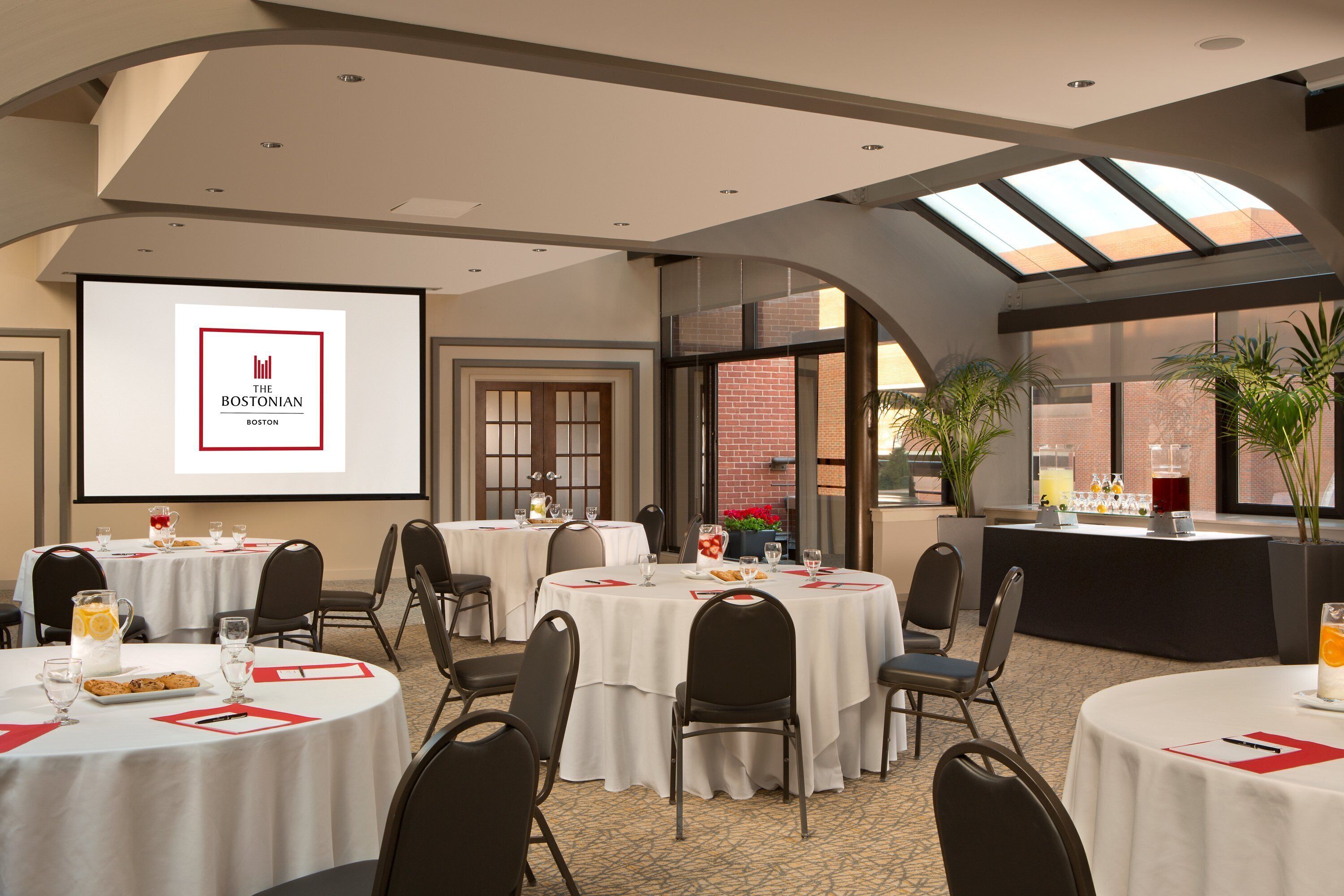 A conference room with multiple tables, chairs, a projector screen displaying 'The Bostonian Boston,' and a bar area with plants and beverages.