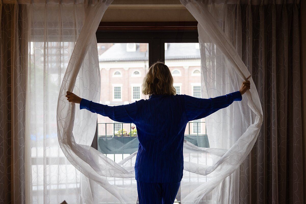 A person standing in a room with large windows, curtains, and a view of buildings outside.