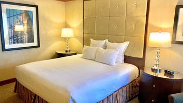 Habitación de hotel con una cama grande, cabecera, mesita de noche con lámpara, reloj y un cuadro en la pared.