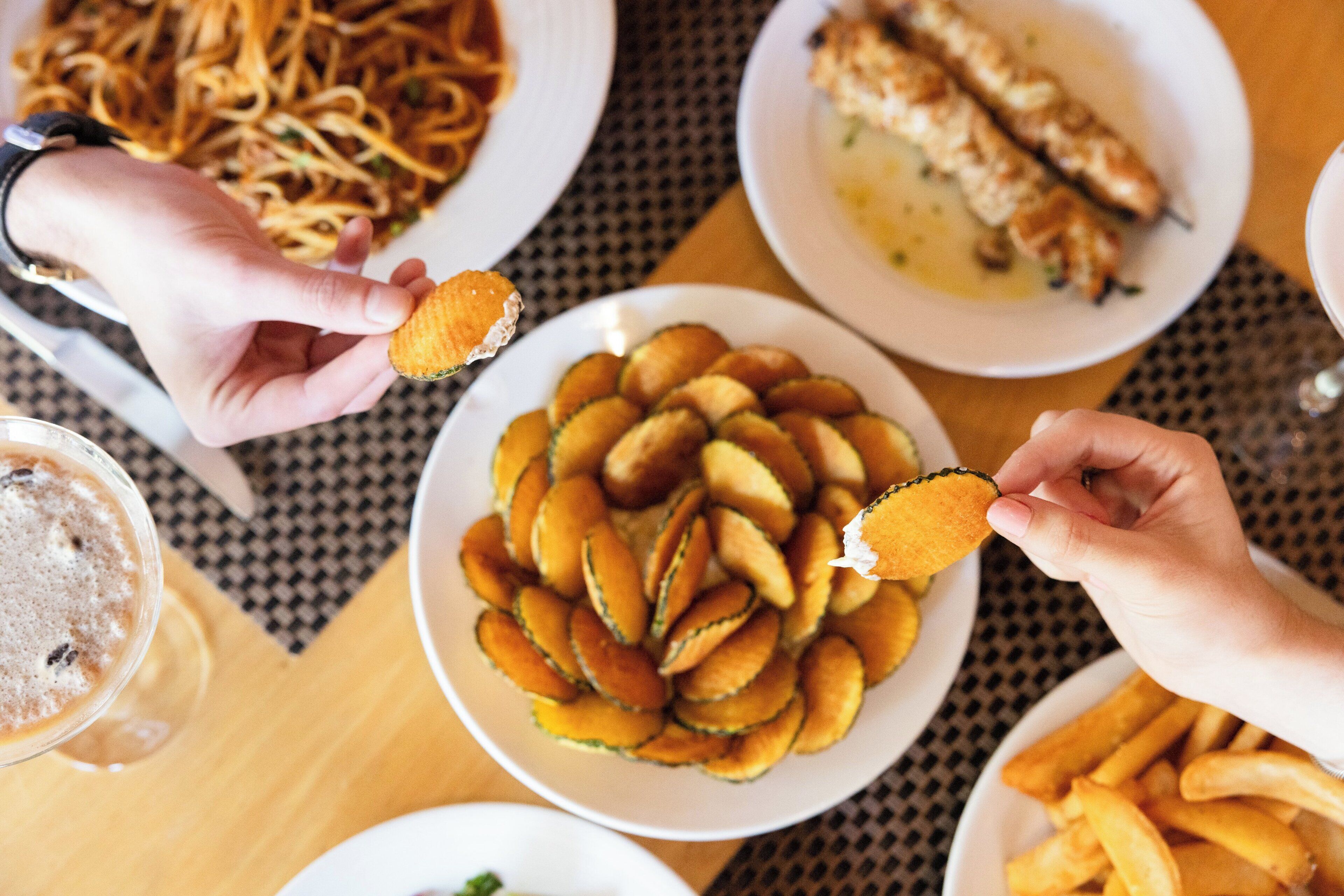 A table with plates of food, a glass of a beverage, and a person's hands holding a food item.