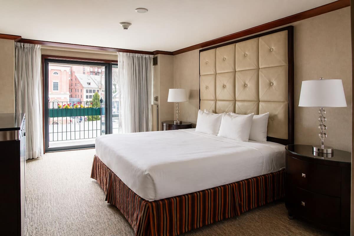 A hotel room with a large bed, a view through a glass door, a bedside table with a lamp, and a dark wooden nightstand.