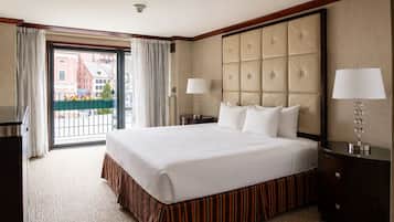 Habitación de hotel con una cama grande, vista a través de un ventanal, una mesita de noche con lámpara y un tocador de madera oscura.
