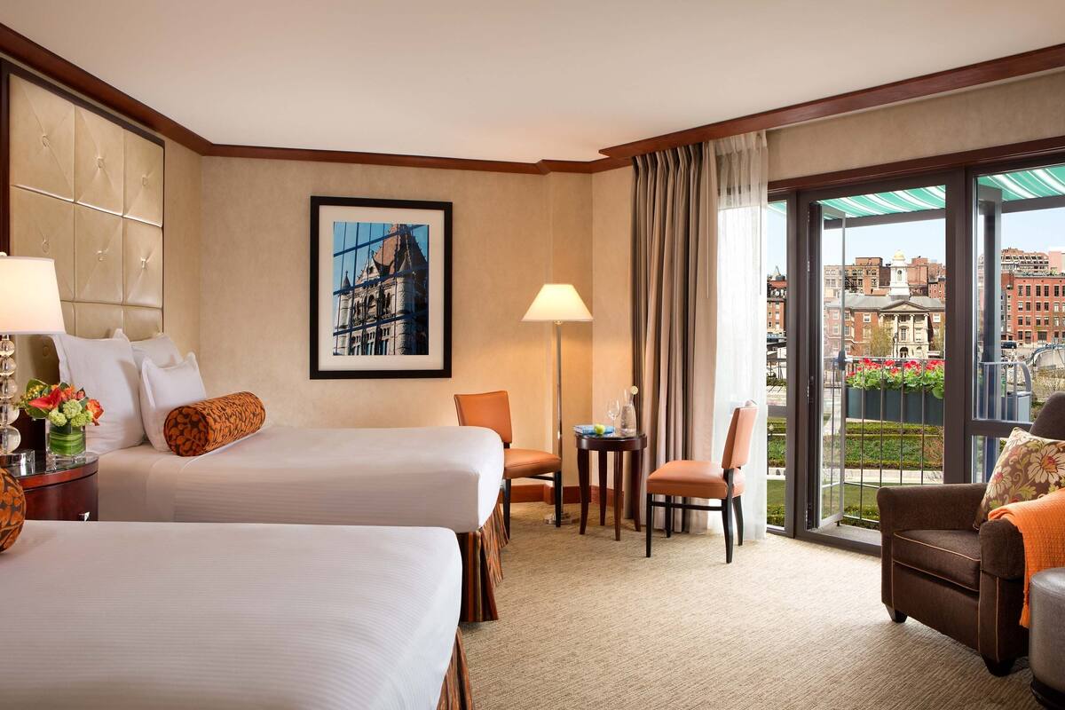 A hotel room with two beds, a desk, a chair, a sofa, a large window with a city view, and a framed picture on the wall.