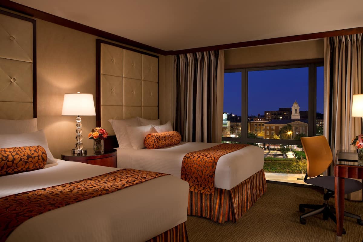 A hotel room with two beds, a desk, a chair, and a large window offering a city view.