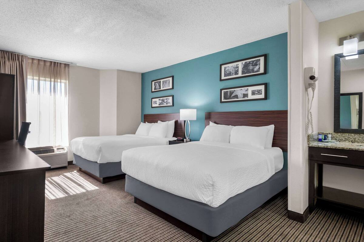 A hotel room with two beds, a desk, a lamp, a mirror, and framed pictures on the wall.