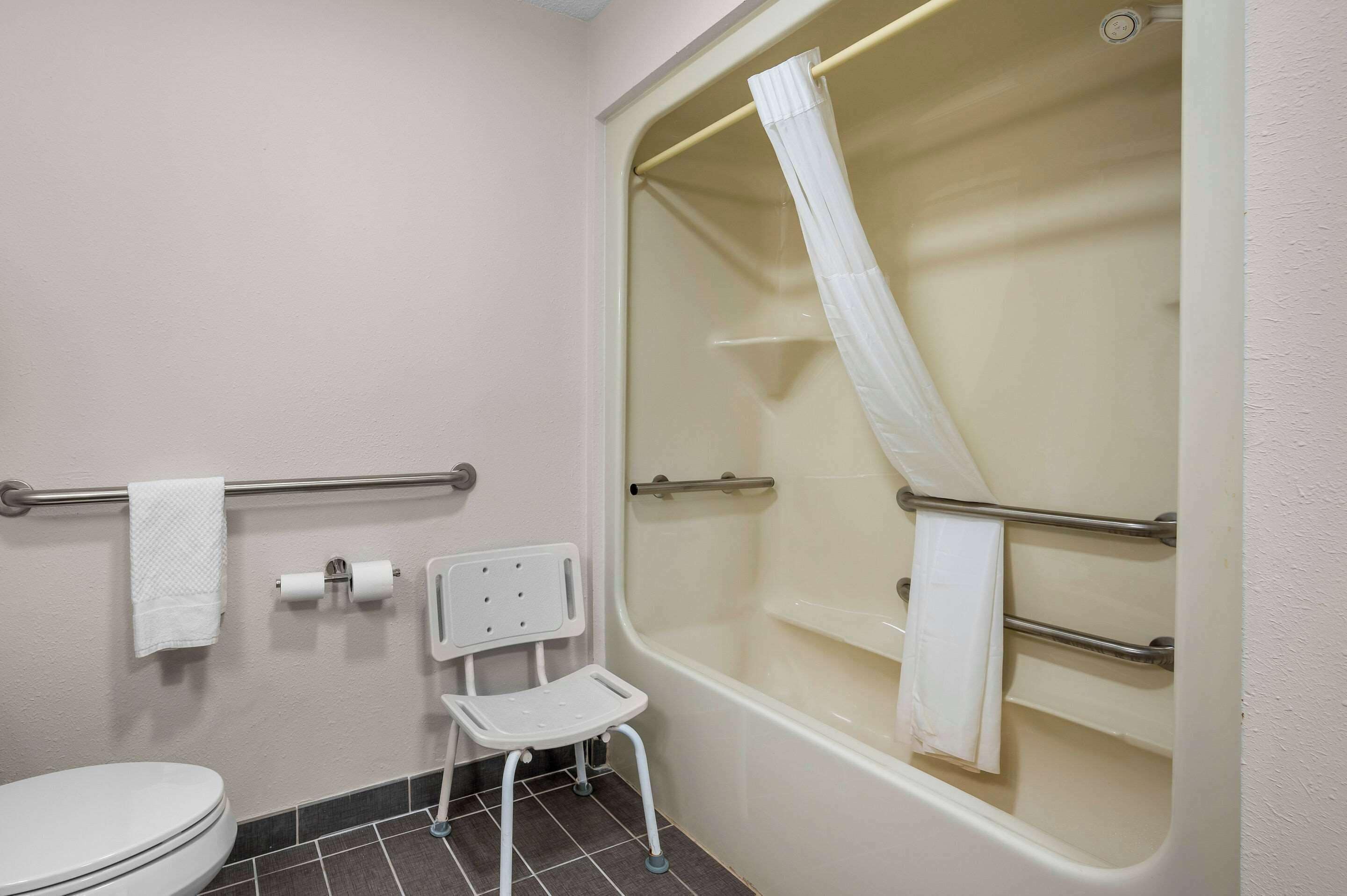 A bathroom with a toilet, a shower with a glass door, a grab bar, a white chair, and a towel rack.