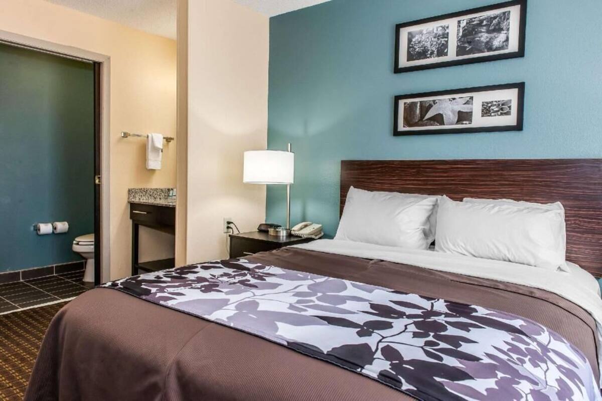 A hotel room with a bed, a nightstand, a lamp, a bathroom with a toilet, and two framed pictures on the wall.