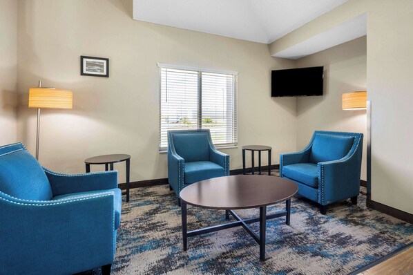 A hotel room with a blue armchair, a round table, a small round table, a floor lamp, a TV mounted on the wall, and a window with blinds.