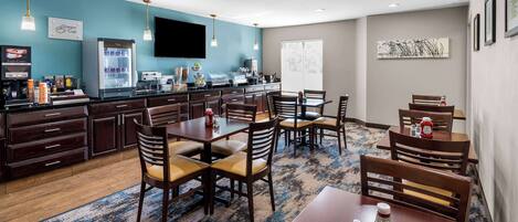 A hotel dining area with multiple tables and chairs, a refrigerator, a coffee station, and a flat-screen TV on the wall.