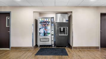 A vending machine and a refrigerator in a hallway with wooden flooring and beige walls.