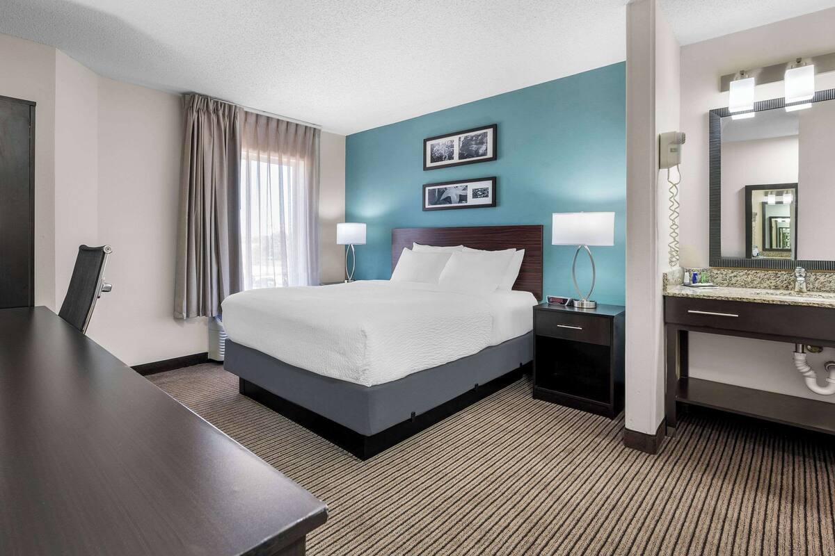A hotel room with a large bed, bedside tables with lamps, a desk, a bathroom with a mirror, and a window with curtains.