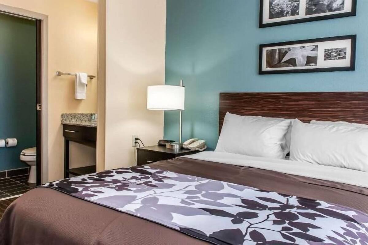 A hotel room with a bed, a nightstand, a lamp, a bathroom with a toilet, and two framed pictures on the wall.