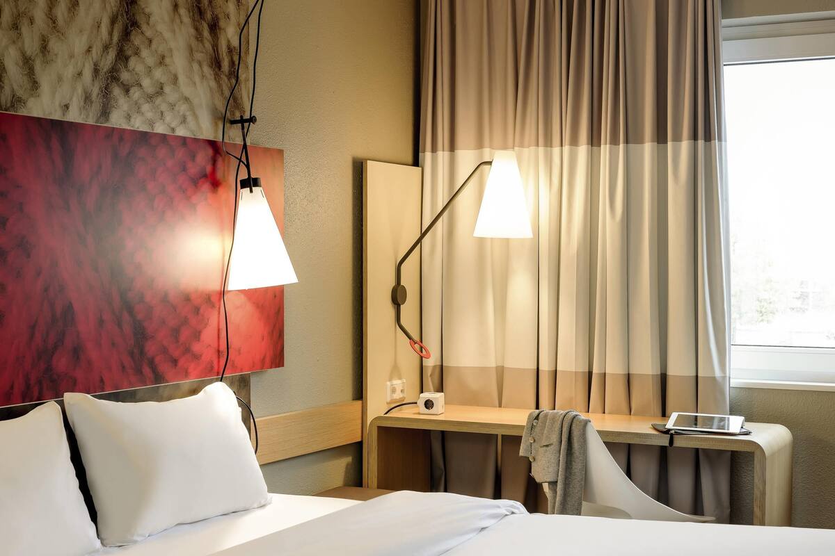 A hotel room with a bed, two bedside lamps, a desk with a laptop, a chair, and a window with curtains.
