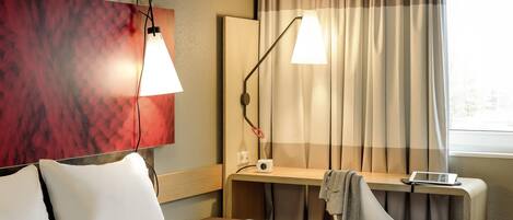 A hotel room with a bed, two bedside lamps, a desk with a laptop, a chair, and a window with curtains.
