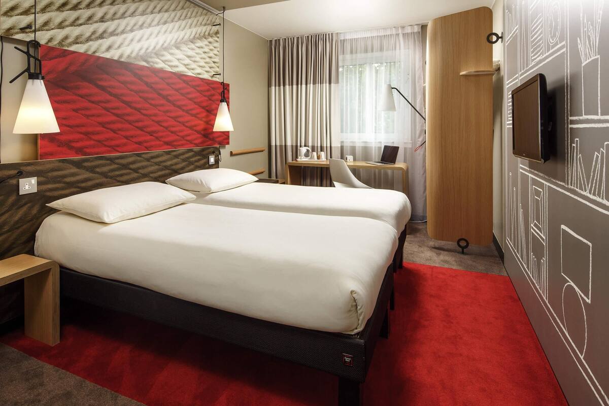 A hotel room with a double bed, a desk with a chair, a TV, and a red carpet.