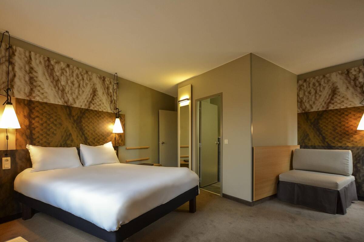 A hotel room with a bed, bedside tables, a chair, and a door leading to a bathroom.