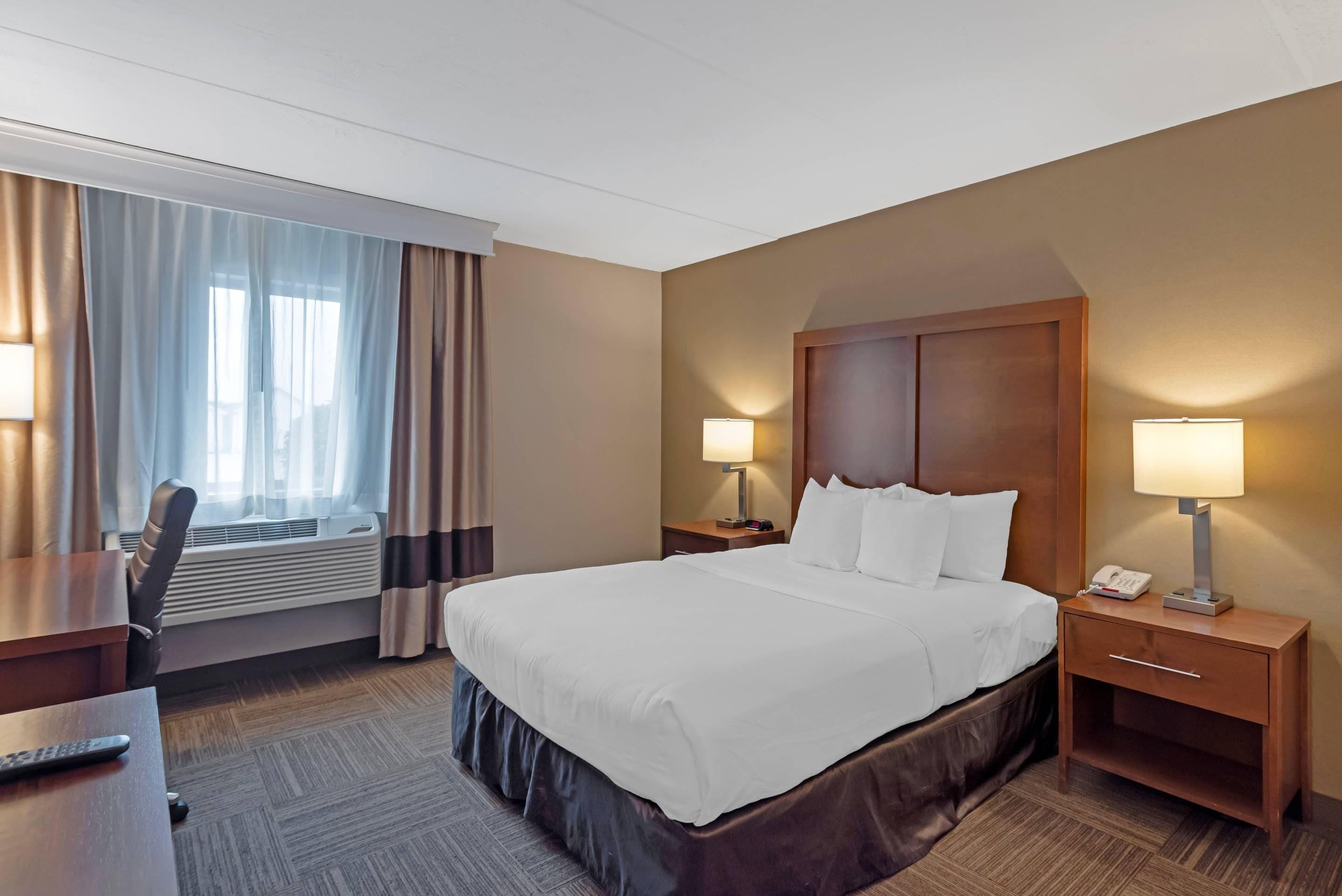 A hotel room with a bed, bedside tables, a desk, an air conditioning unit, and curtains.
