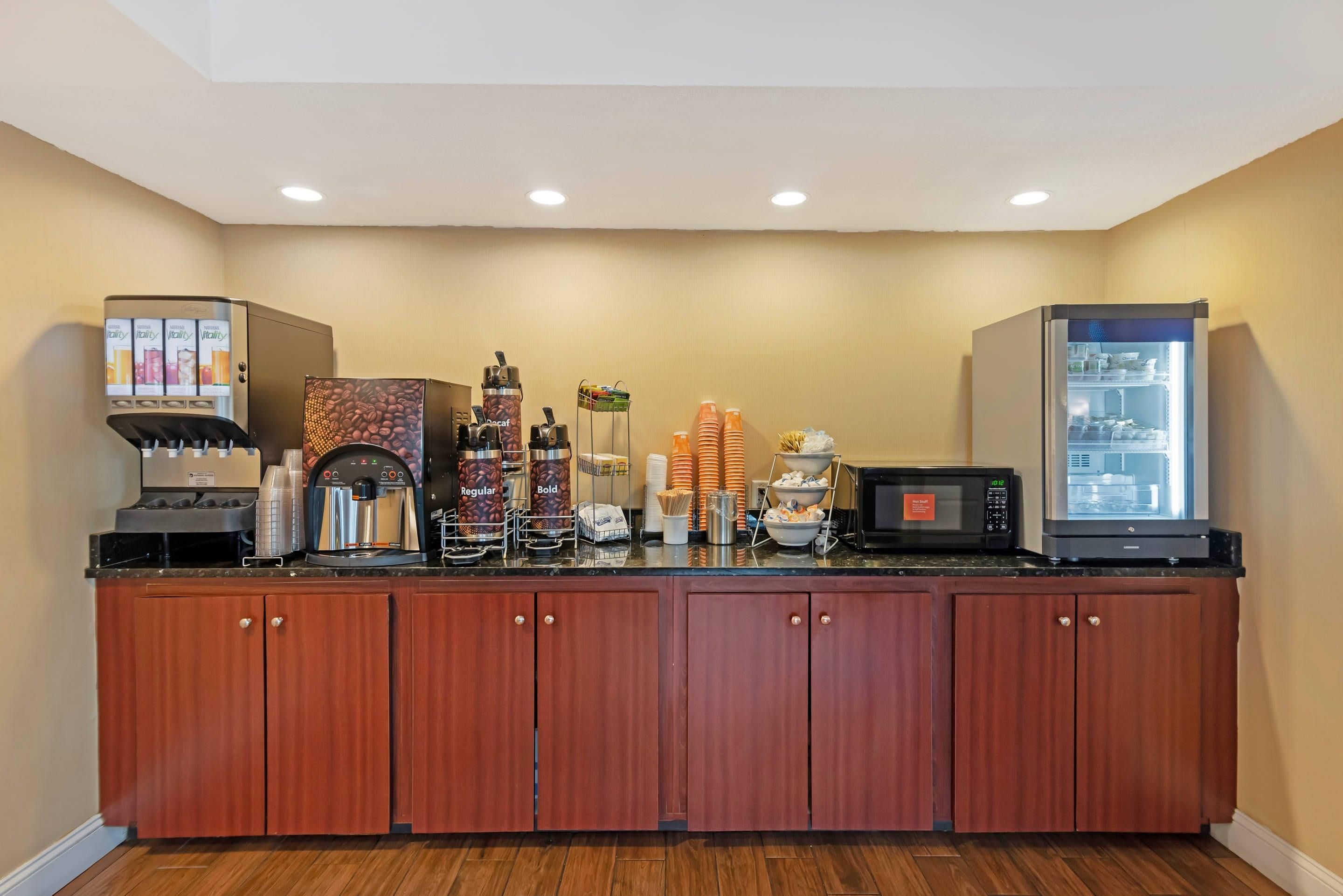 A hotel breakfast area with a coffee machine, a refrigerator, and various food items.