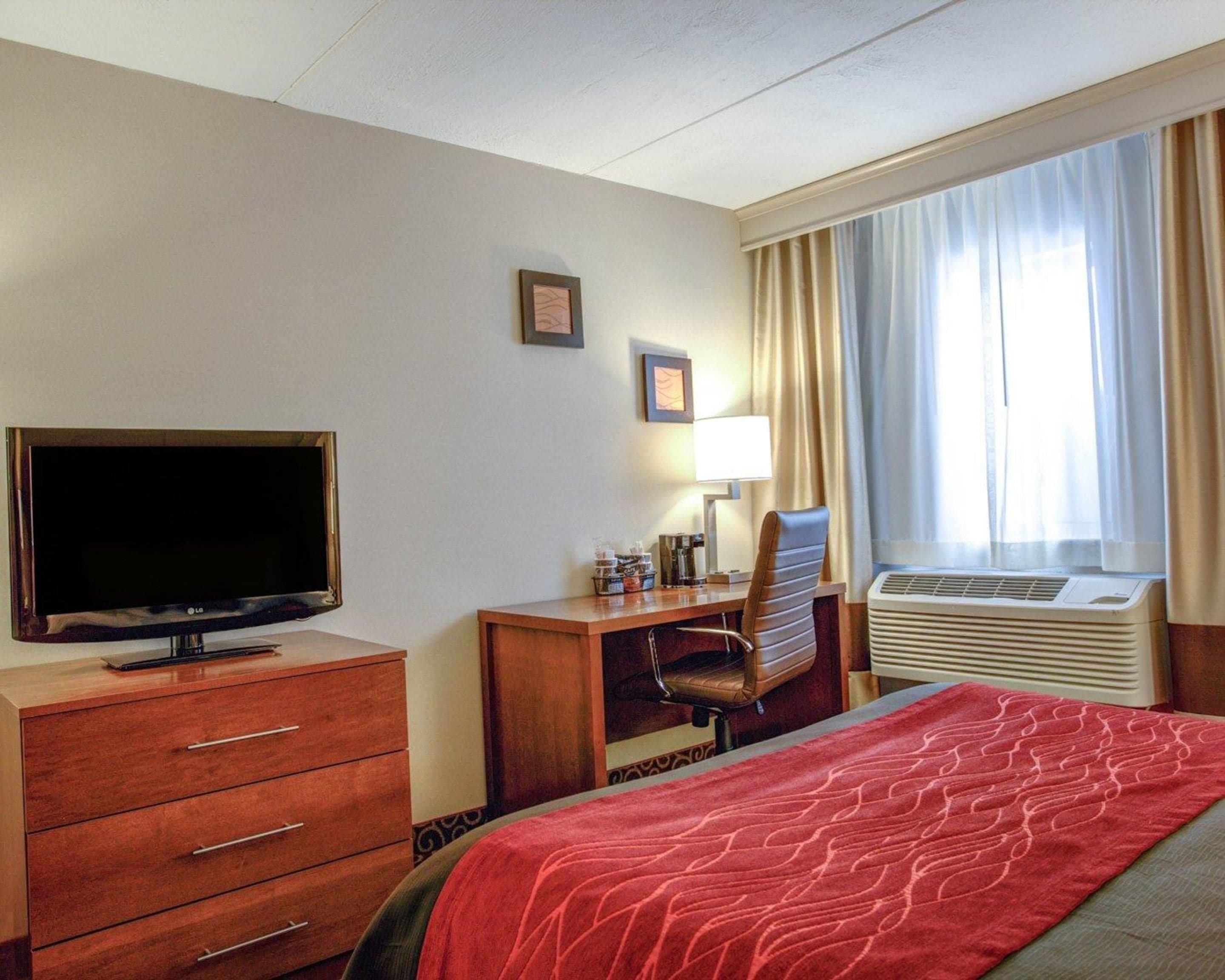 A hotel room with a bed, a desk with a chair, a television, a lamp, and an air conditioning unit.