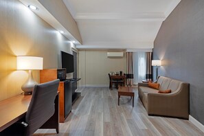 Habitación de hotel con televisor de pantalla plana, escritorio con lámpara, sofá, mesa de comedor con sillas y unidad de aire acondicionado.