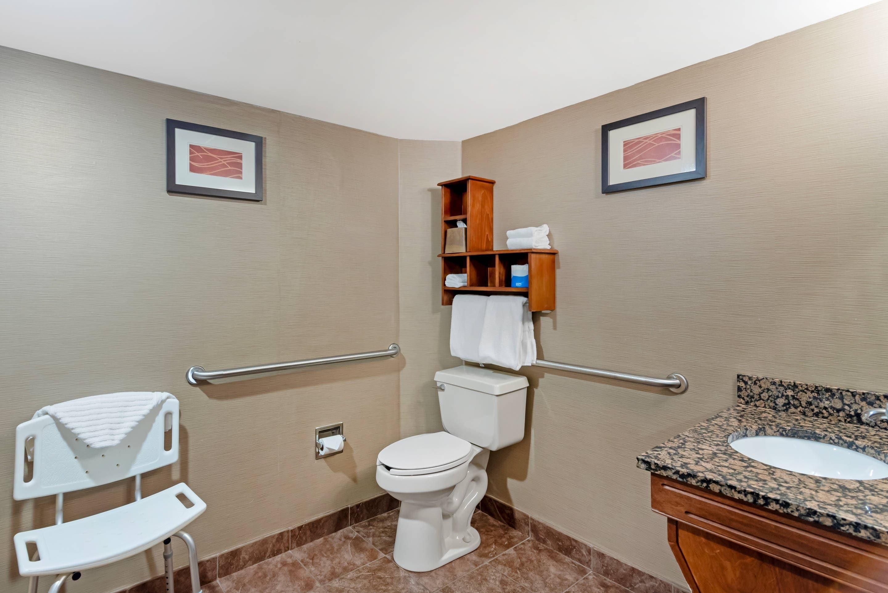 A bathroom with a toilet, a sink, a handrail, a white chair, and two framed pictures on the wall.