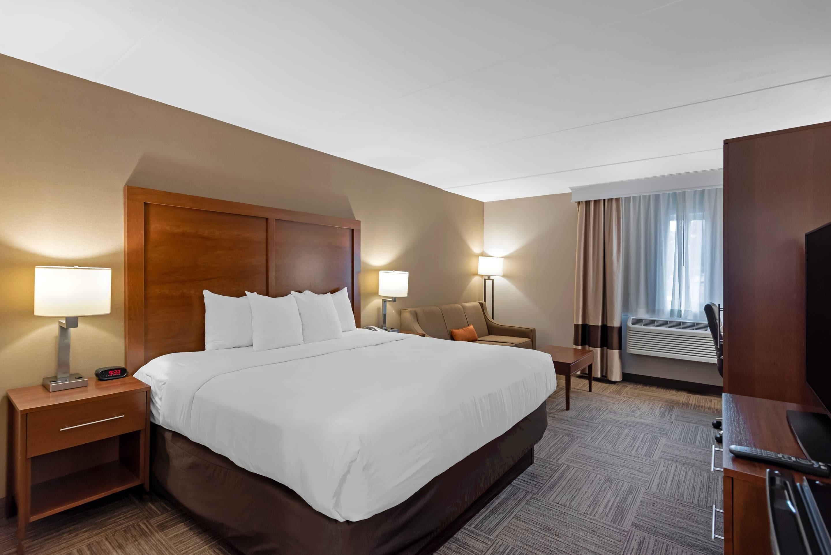 A hotel room with a large bed, bedside lamps, a desk, a chair, an air conditioning unit, and a television.