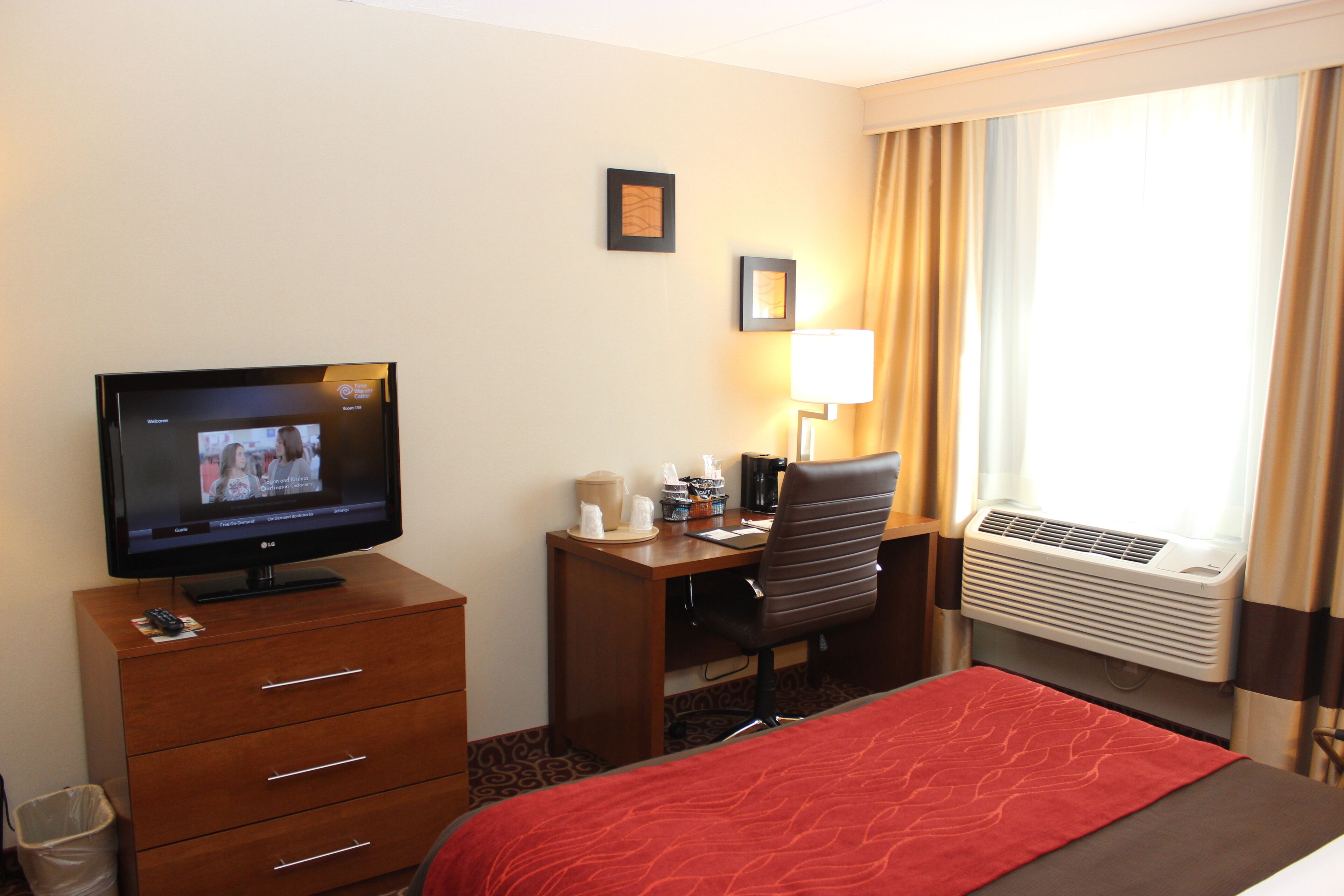 A hotel room with a bed, a desk with a chair, a television, a lamp, and an air conditioning unit.