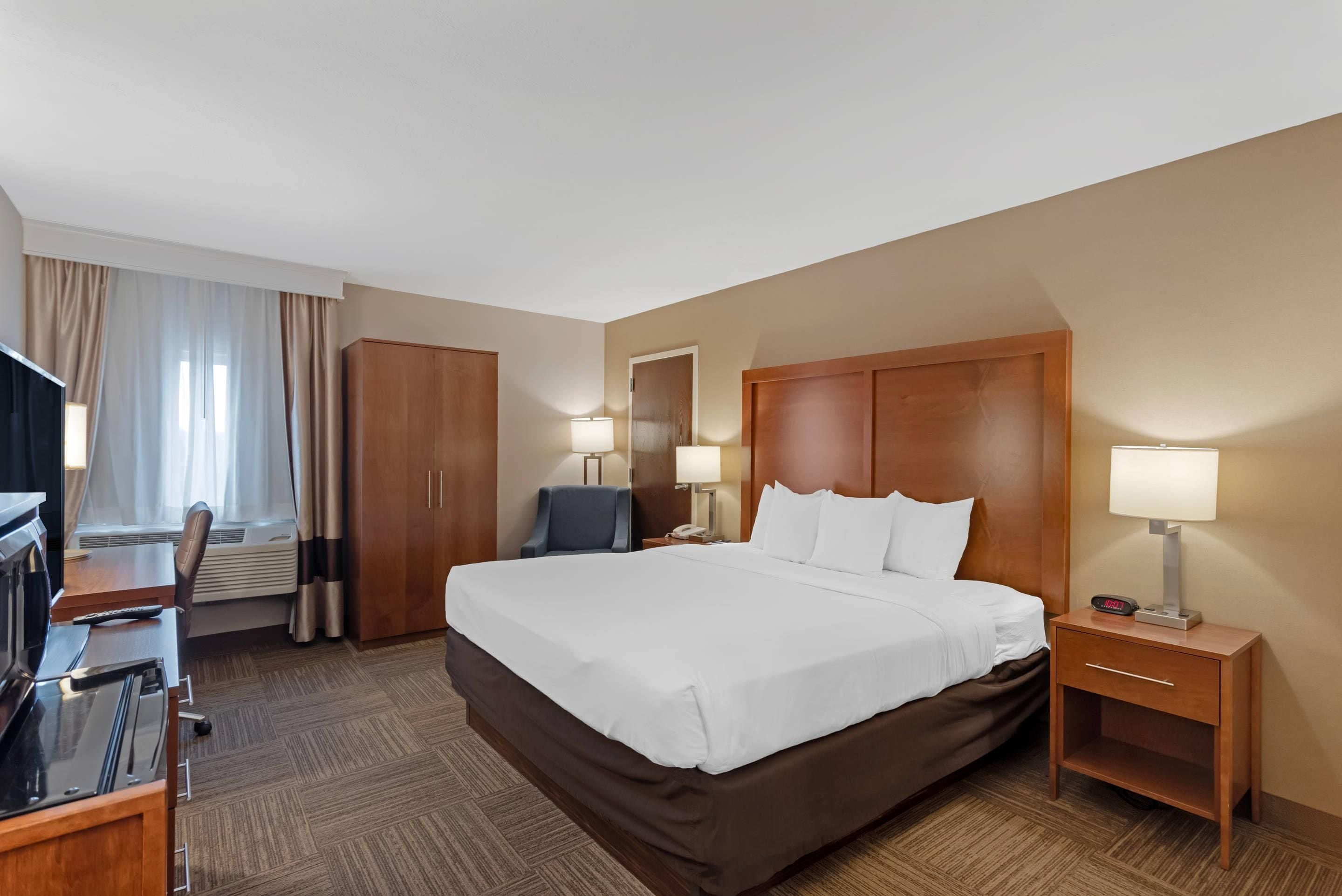 A hotel room with a large bed, a desk with a chair, a TV, and a bedside table with a lamp.