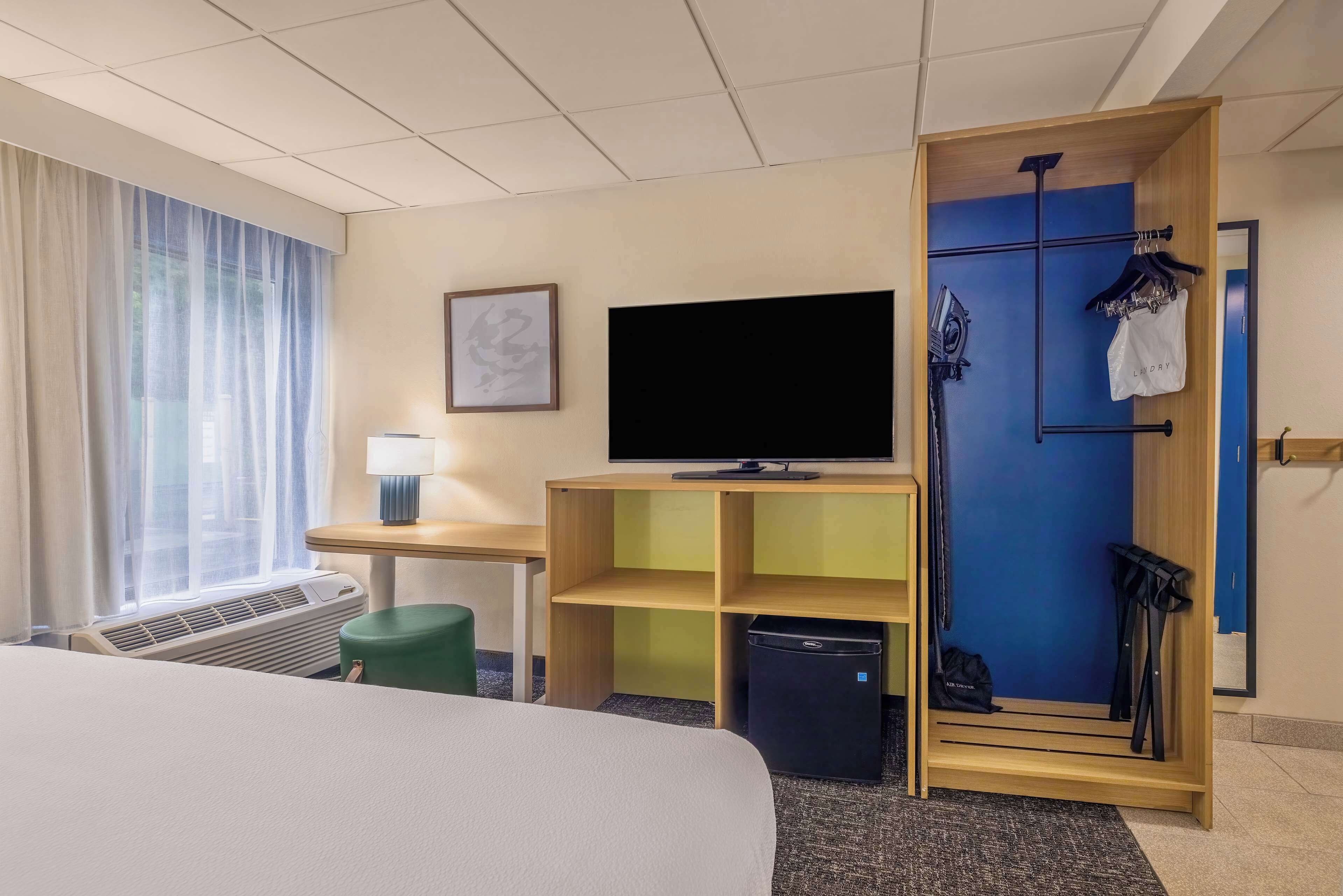 Habitación de hotel con cama, televisión, escritorio, silla, pequeña mesa, lámpara, armario y ventana con cortinas.
