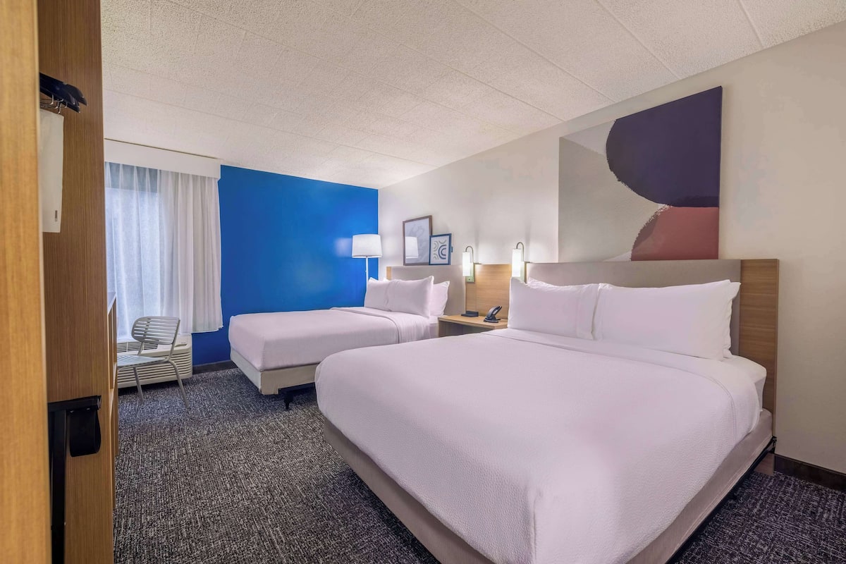 Une chambre d’hôtel avec deux lits, un bureau, une chaise, une fenêtre avec des rideaux et un mur orné d’un tableau.