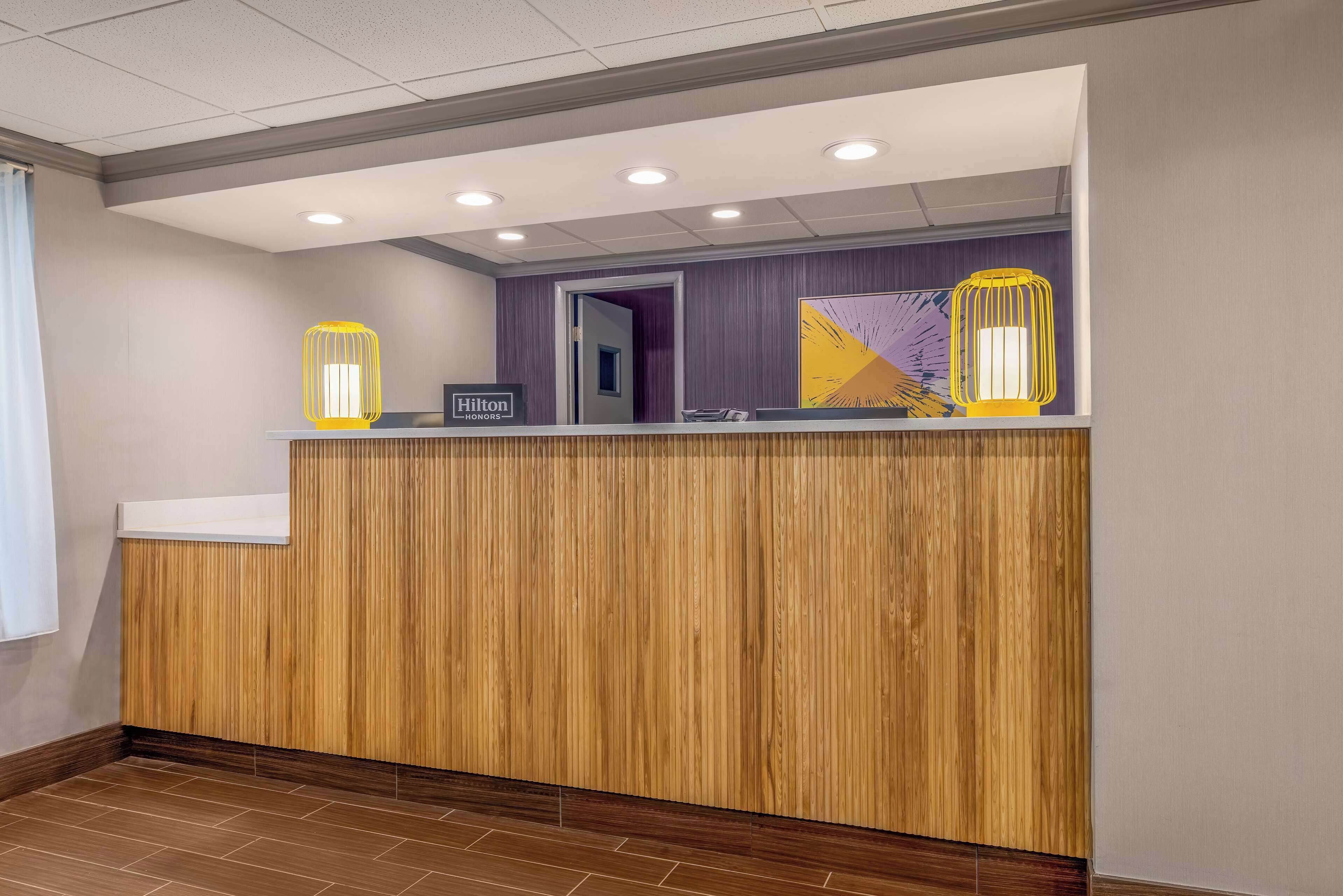 A hotel reception area with a wooden reception desk, a "Hilton Honors" sign, and two yellow wall-mounted lamps.
