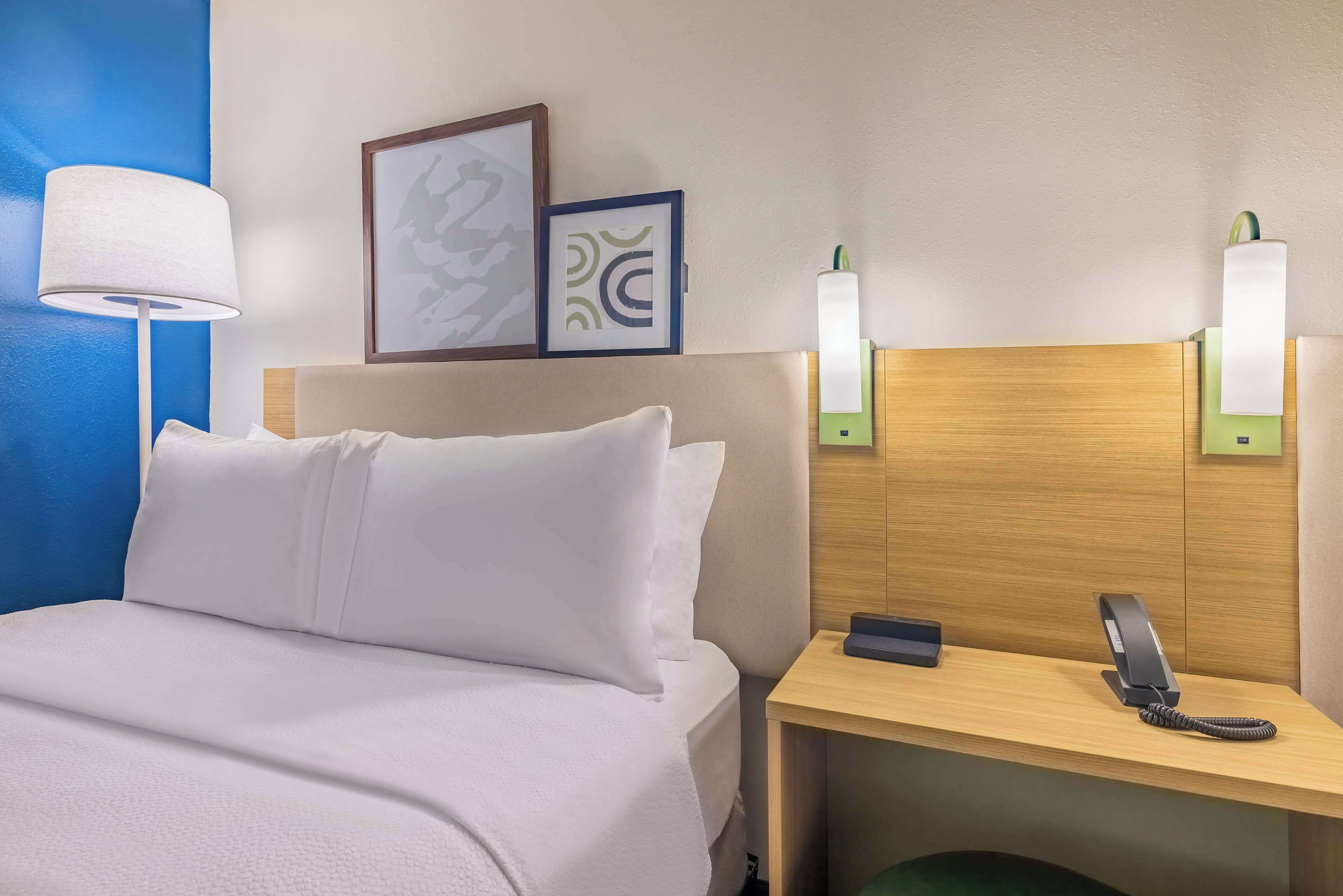 A hotel room with a bed, bedside lamp, wall art, and a desk with a phone.