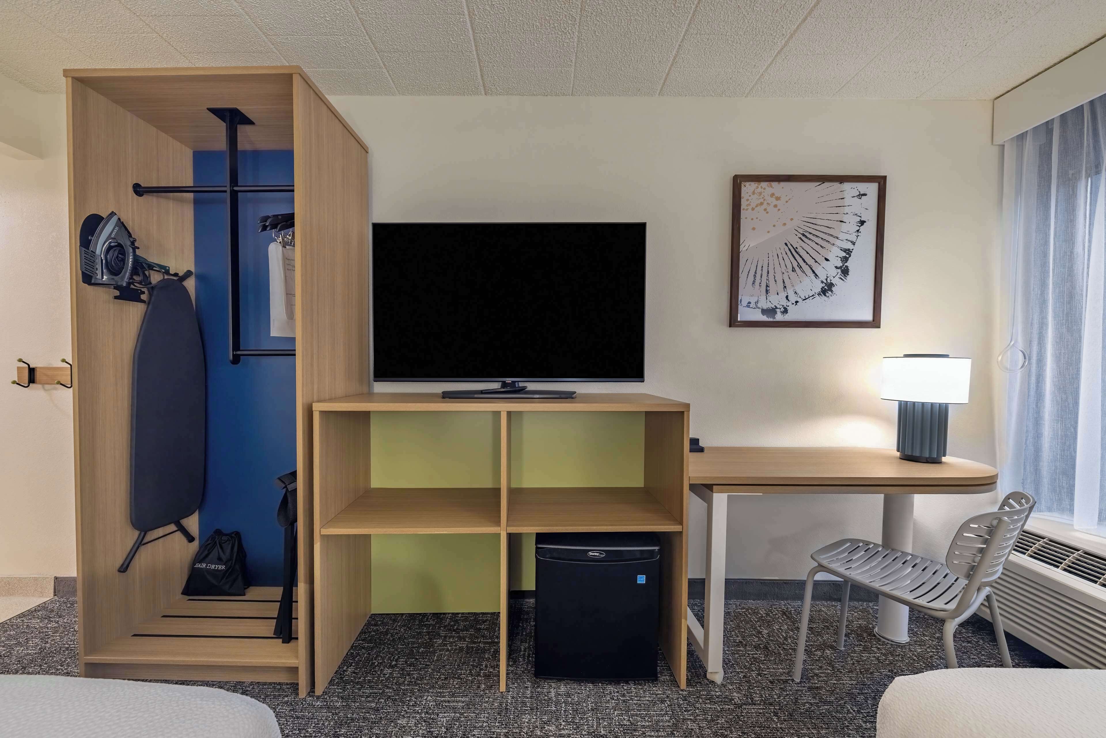 Habitación de hotel con cama, escritorio con lámpara, televisor, mini refrigerador, silla, armario y un cuadro en la pared.