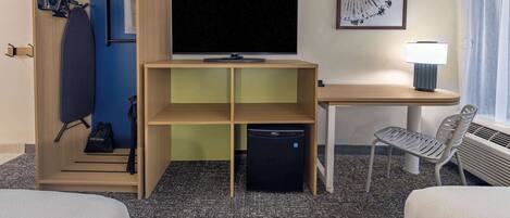 A hotel room with a bed, a desk with a lamp, a TV, a mini-fridge, a chair, a wardrobe, and a framed picture on the wall.