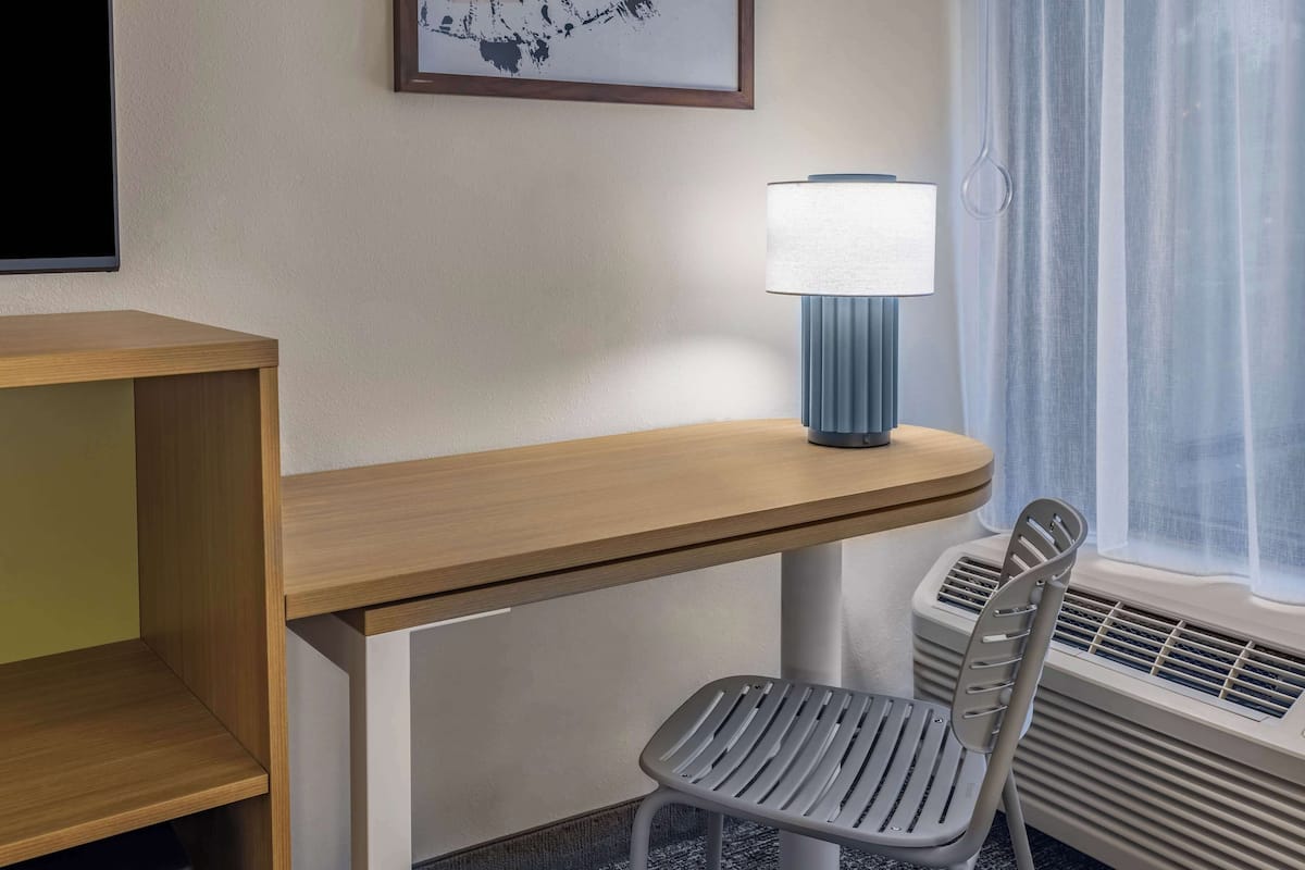 Une chambre d’hôtel avec un bureau en bois, une chaise grise, une lampe fixée au mur et une fenêtre avec des rideaux.