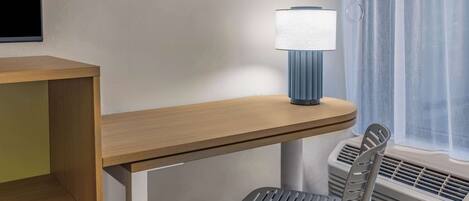 A hotel room with a wooden desk, a grey chair, a wall-mounted lamp, and a window with curtains.