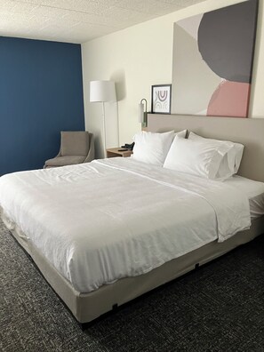 A hotel room with a large bed, a chair, a nightstand with a lamp, and a wall with abstract art.