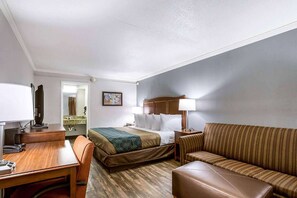 A hotel room with a bed, a desk, a chair, a sofa, and a nightstand with a lamp.