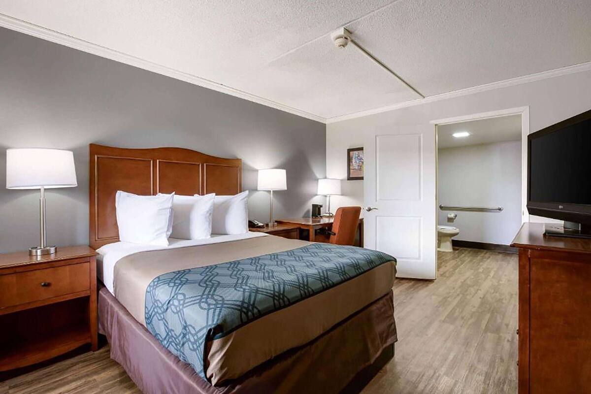 A hotel room with a large bed, two bedside tables with lamps, a desk with a chair, a television, and a bathroom visible through an open door.