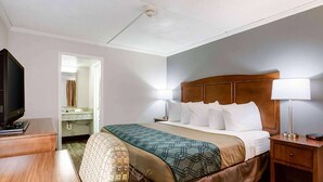 A hotel room with a bed, bedside lamps, a TV, a desk, and a bathroom visible through an open door. - Travelodge by Wyndham Canton/Livonia Area, MI (Canton)